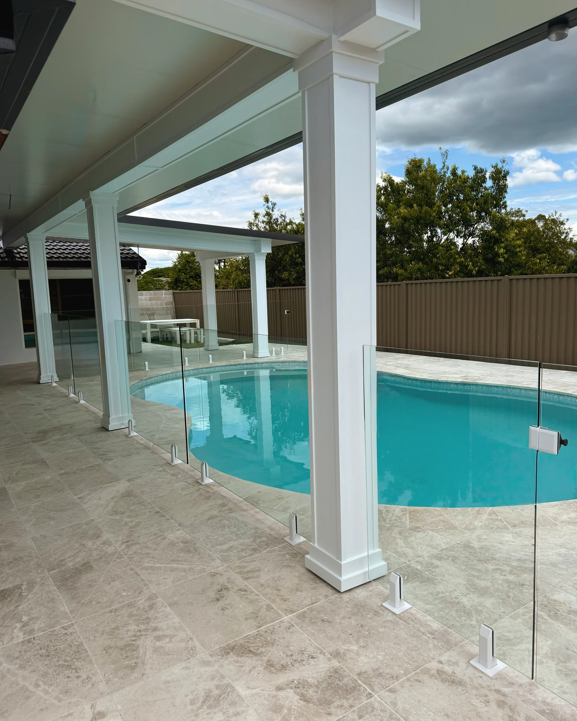 Pale marble-look pavers around a curved pool behind a frameless glass fence